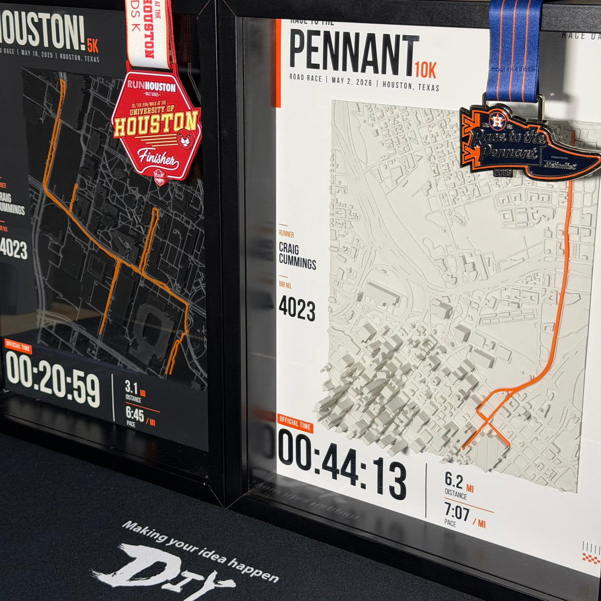 Two framed marathon or 10K race maps displayed on a desk, one for Houston and one for Pennant, each with a ribbon and medal, in a real indoor setting.