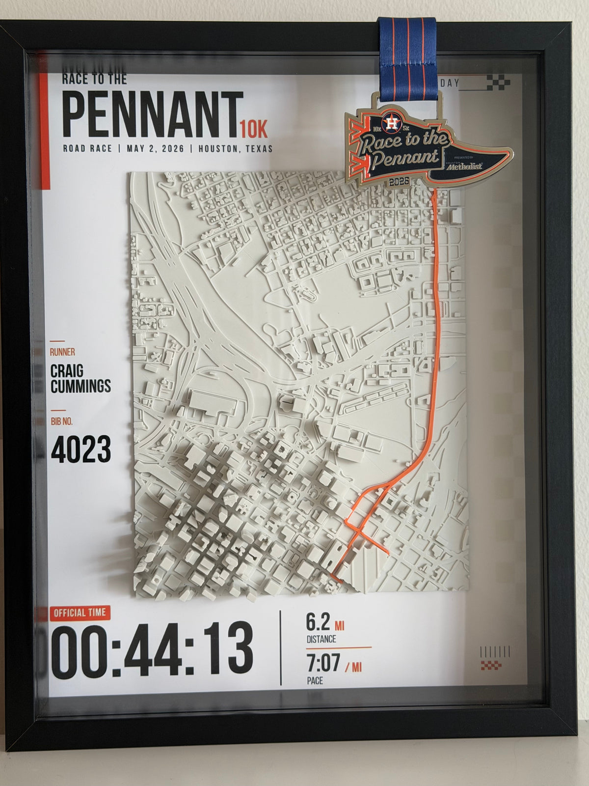 A framed 3D papercity wall art piece celebrating the 2026 Pennant Road Race. The black-framed display features a detailed beige papercity map with an orange route line, a blue and orange ribbon, a commemorative race pin, and printed race details including the name Craig Cummings, number 4023, and official time 00:44:13.